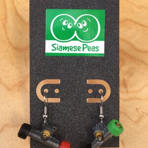 May include: A pair of novelty earrings made from miniature skateboard parts. The earrings feature silver-colored metal trucks with black, red, and green wheels. The earrings are displayed on a black card with the text "Siamese Peas".