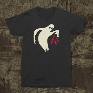 May include: Dark gray t-shirt featuring a cream-colored ghost design with red lightning bolts emanating from its hand. The ghost has a simple, minimalist design. The shirt is made of a soft material.