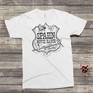 Op de afbeelding: Wit T-shirt met een zwart-witte afbeelding van een filmranch-bord met de tekst "Spahn Movie Ranch Come Join the Family!"
