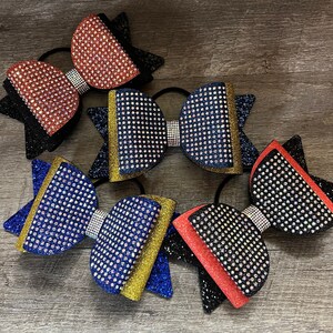 May include: Several rhinestone-studded hair bows in various colors, including red, black, and blue. Each bow features a glittery accent and a rhinestone-covered center. The bows are arranged on a wooden surface.