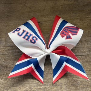 Op de afbeelding: Een witte, rode en blauwe cheerleading strik met de tekst "PJHS" in blauw. De strik heeft een chevron ontwerp en een schoollogo. De strik is gemaakt van een lintmateriaal.
