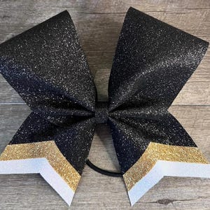 Op de afbeelding: Een grote haarstrik met een zwarte glitterafwerking en een chevronpatroon aan de onderkant. Het chevronontwerp bestaat uit een gouden glitterstreep gevolgd door een witte glitterstreep. De strik is bevestigd met een zwart elastiekje.