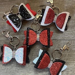 Puede incluir: Seis llaveros de lazo de brillo rojo, negro y blanco con diamantes de imitación. Los lazos están superpuestos y tienen una franja negra en el medio.