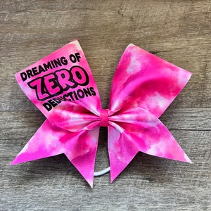 May include: A pink and white tie-dye cheer bow with the text "Dreaming of Zero Deductions" in black.