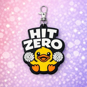 May include: A black rubber keychain featuring a yellow duck character holding white pom-poms. The text HIT ZERO is printed in bold white letters above the duck. The keychain includes a silver-colored metal lobster clasp at the top for attachment.