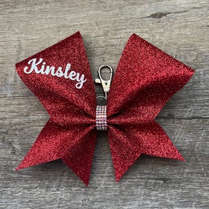 May include: Red glitter cheer bow with a silver clip and a rhinestone embellishment. The bow is personalized with the name "Kinsley" in white.