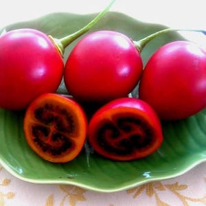 May include: Three whole bright red tamarillo fruits with green stems and two halved tamarillo fruits showing orange flesh with dark seeds, all arranged on a green ceramic dish shaped like a large leaf with a textured surface.