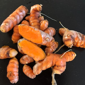 May include: A group of turmeric root, some of which are cut open to reveal the bright orange flesh. The roots are a vibrant orange and yellow color and have a rough, knobby texture.