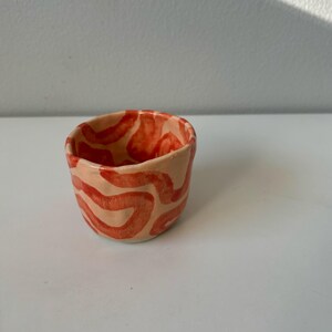May include: A small, hand-crafted ceramic cup with a light beige glaze and a red abstract pattern.
