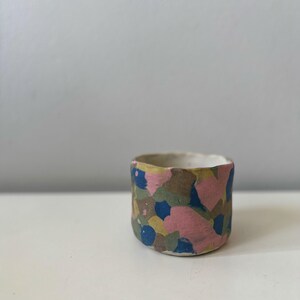 May include: A small, handmade ceramic cup with a colourful, abstract pattern. The exterior features a mosaic of pink, blue, green, and yellow shapes. The interior is a plain, off-white colour. The cup is approximately 7.6 cm wide and 6.4 cm tall.