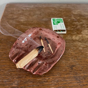 Op de afbeelding: Een rode keramische wierookhouder met zwarte stippen en een brandende wierookstok van hout erin. Drie lucifers zitten ook in de houder. Een doosje lucifers met een groen boomlogo en de tekst "borderley" en "de natuur" ligt op de houten tafel.