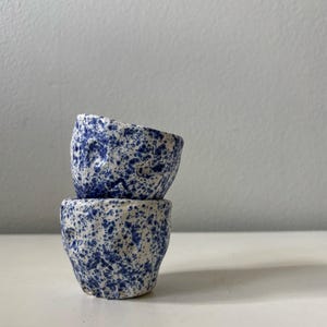 Könnte beinhalten: Zwei kleine, handgefertigte Keramikbecher, die übereinander gestapelt sind. Die Becher sind cremefarben mit einem gesprenkelten blauen Muster. Sie haben eine raue, strukturierte Oberfläche und eine unregelmäßige Form, was darauf hindeutet, dass sie handgefertigt sind.