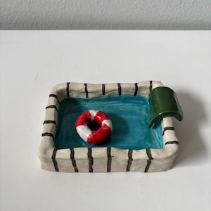 Könnte beinhalten: Ein keramischer Miniaturpool mit einem rot-weißen Rettungsring, der in blauem Wasser schwimmt. Der Pool hat einen beigen Rand mit schwarzen Streifen und ein grünes Sprungbrett.