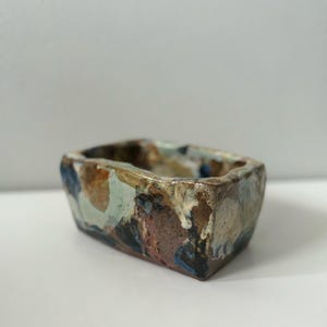 May include: A small, rectangular ceramic planter with a brown, white, blue and green glaze. The planter has an irregular shape and a rough texture.