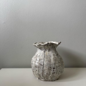May include: A textured, off-white ceramic vase with a ruffled rim. The vase has vertical grooves and dark grey accents, giving it a rustic appearance. The vase is sitting on a white surface.