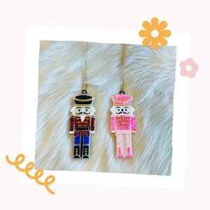 May include: Two wooden nutcracker ornaments, one in red and blue with gold accents and the other in pink with gold accents. Both have a white background and glitter details.