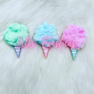 May include: Three cotton candy cookies decorated with pink, blue, and green frosting. The cookies are shaped like cones and are sitting on a white fluffy surface. The text "Lil's Freshies" is written in pink neon letters across the cookies.