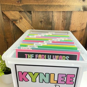 Op de afbeelding: Een doorzichtige plastic opbergbak gevuld met kleurrijke ordners. De bak heeft een label met de tekst "KYNLEE Ray" in een speels lettertype. De ordners zijn gelabeld met klasniveaus en "THE EARLY YEARS".