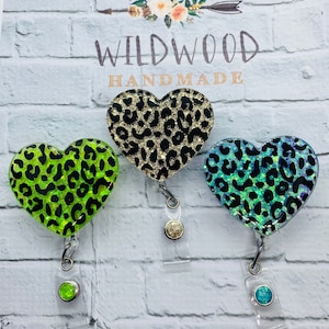 May include: Three heart-shaped retractable badge holders with leopard print designs. One is neon green, one is gold and black, and the third is blue and green. The text "Wildwood Handmade" is above the badge holders.