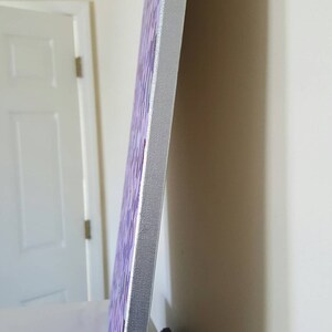 May include: A close-up of the side of a canvas painting. The canvas is wrapped around a wooden frame and has a purple and white abstract design. The edges of the canvas are painted white.