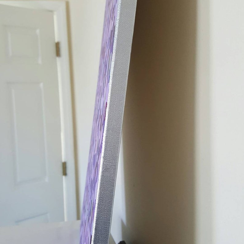 May include: A close-up of the side of a canvas painting. The canvas is wrapped around a wooden frame and has a purple and white abstract design. The edges of the canvas are painted white.