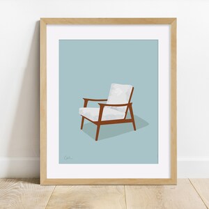 May include: A framed print featuring a mid-century modern armchair. The chair has a white cushion and backrest, with brown wooden arms and legs. The background is a light blue, and the frame is light brown.