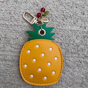 May include: A yellow pineapple-shaped keychain with white dot details and a green top. The keychain has a gold clasp and a charm with the word "Starr". Decorative red beads and green leaves are attached.