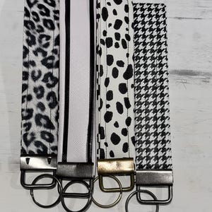 May include: Four fabric wristlet keychains featuring distinct black and white patterns: leopard print, a white stripe with black borders, dalmatian spots, and a houndstooth check. Each strap is secured with a metal hardware clasp and a circular ring.