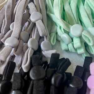 May include: A close-up of a variety of colorful elastic cords with plastic toggles. The cords are in shades of gray, light green, black, and pink.