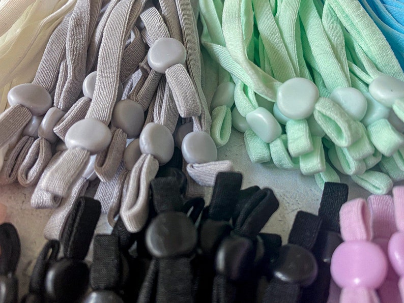 May include: A close-up of a variety of colorful elastic cords with plastic toggles. The cords are in shades of gray, light green, black, and pink.