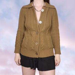 May include: A tan cable-knit cardigan with a collared neckline and large, round buttons. The long-sleeved sweater features a textured cable knit design on the front and sleeves. The cardigan is buttoned up and appears to be made of a soft, warm material.