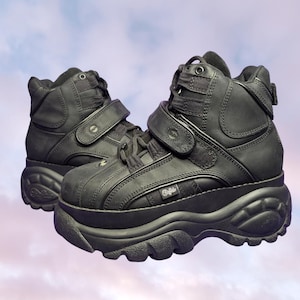 May include: Pair of black platform boots with thick soles, featuring adjustable straps and laces. The boots have a high-top design and a textured, chunky sole. The brand name "Buffalo" is visible on the side.