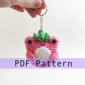 May include: A pink crocheted frog with a green crocheted leaf on its head. The frog is holding a white crocheted ball. The frog is attached to a silver key chain.