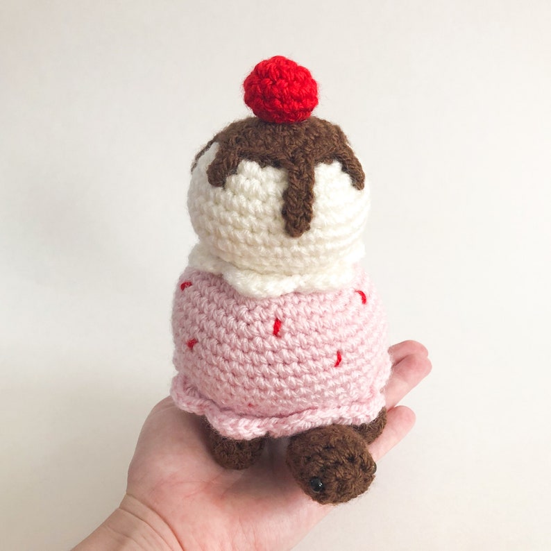 May include: A crocheted ice cream cone toy with a pink cone, white ice cream, chocolate sauce, and a red cherry on top.