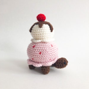May include: A crocheted turtle wearing a pink and white ice cream cone with a red cherry on top.