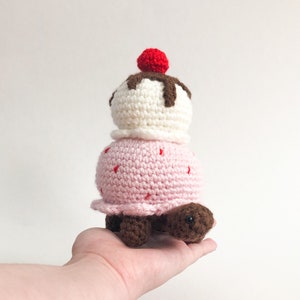 May include: A crocheted turtle toy shaped like an ice cream cone. The turtle is pink with a white ice cream scoop on top, topped with a red cherry and brown chocolate sauce.
