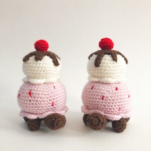 May include: Two crocheted turtle toys shaped like ice cream cones. Each turtle has a pink ice cream cone body with a white ice cream scoop and a red cherry on top. The ice cream scoop has brown chocolate sauce drizzled over it.