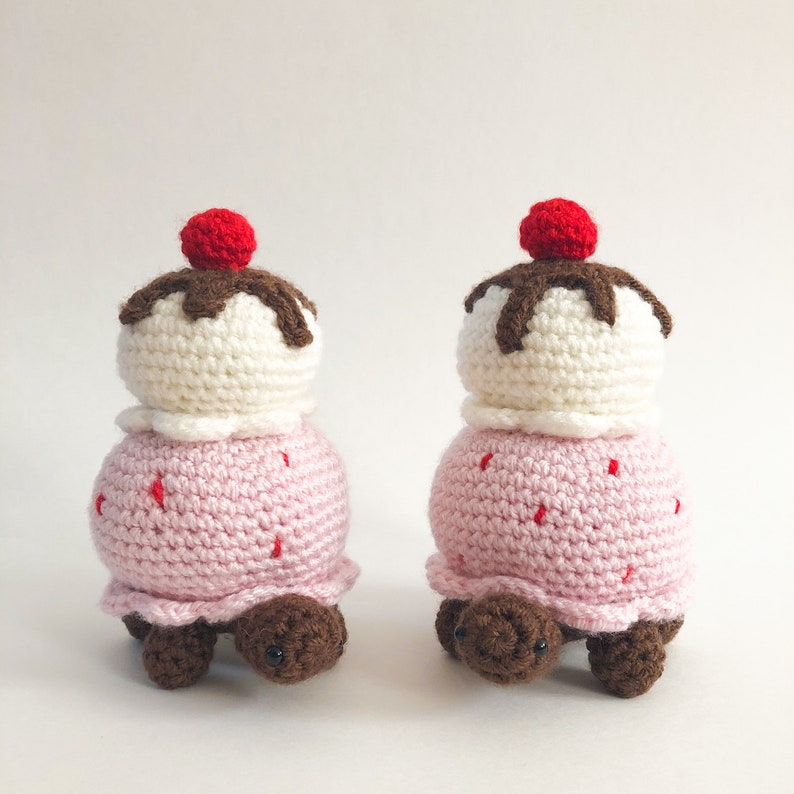 May include: Two crocheted turtle toys shaped like ice cream cones. Each turtle has a pink ice cream cone body with a white ice cream scoop and a red cherry on top. The ice cream scoop has brown chocolate sauce drizzled over it.