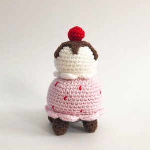 May include: A crocheted ice cream cone toy with a pink base, white ice cream, brown chocolate topping, and a red cherry on top. The toy has four brown legs.