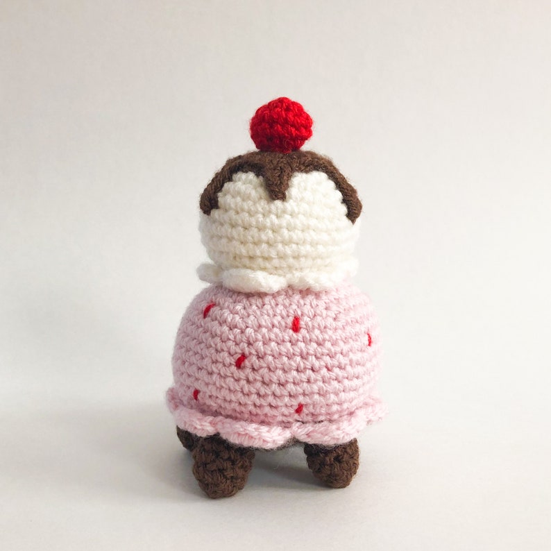 May include: A crocheted ice cream cone toy with a pink base, white ice cream, brown chocolate topping, and a red cherry on top. The toy has four brown legs.
