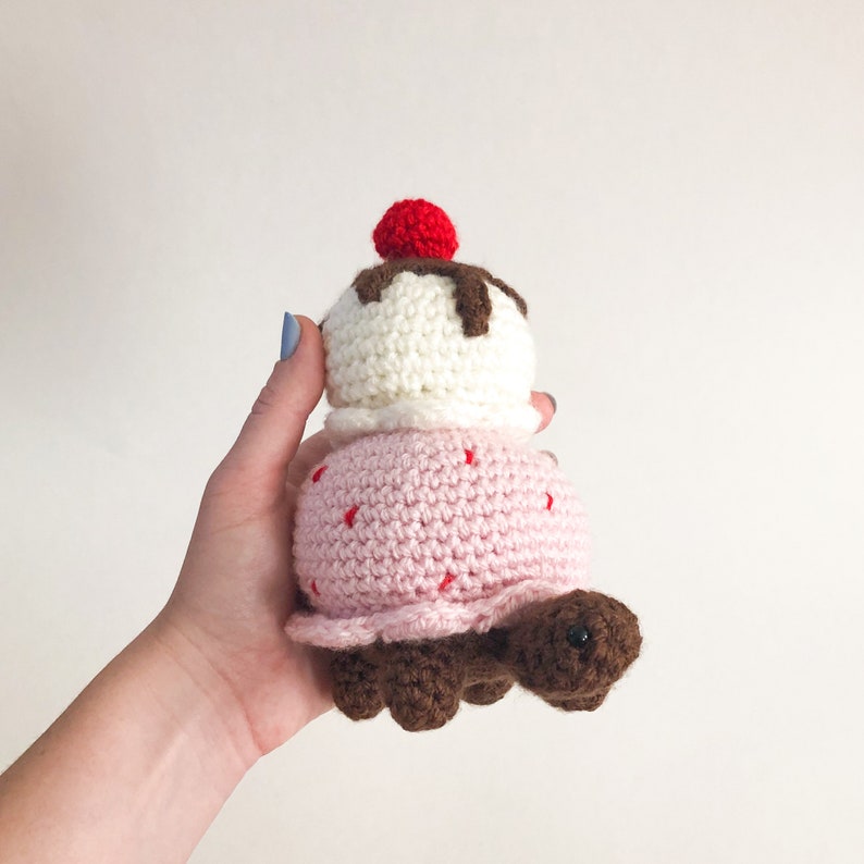 May include: A crocheted ice cream cone turtle toy with a pink shell, white ice cream, brown chocolate sauce, and a red cherry on top.