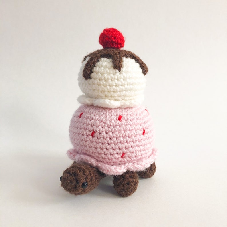 May include: A crocheted turtle wearing a pink and white ice cream cone with a red cherry on top.
