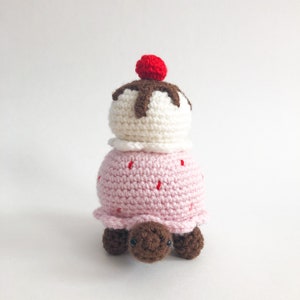 May include: A crocheted turtle with a pink and white ice cream cone on its back. The ice cream cone has a red cherry on top and brown chocolate sauce.