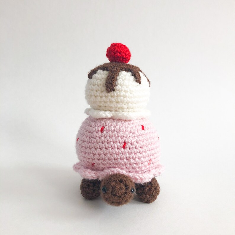May include: A crocheted turtle with a pink and white ice cream cone on its back. The ice cream cone has a red cherry on top and brown chocolate sauce.