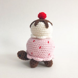 May include: A crocheted turtle toy shaped like an ice cream cone. The turtle is pink with red sprinkles and a white ice cream scoop with brown chocolate topping and a red cherry on top.