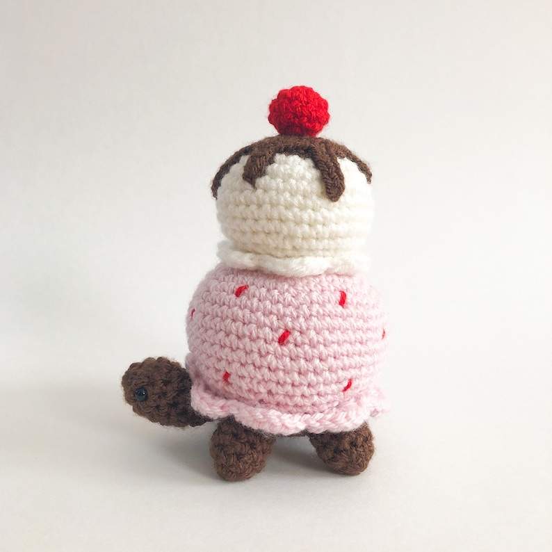 May include: A crocheted turtle toy shaped like an ice cream cone. The turtle is pink with red sprinkles and a white ice cream scoop with brown chocolate topping and a red cherry on top.