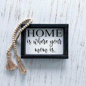May include: A black framed sign with white background and the text "HOME is where your mom is." The sign is on a white wood background with a wooden bead garland with tassels.