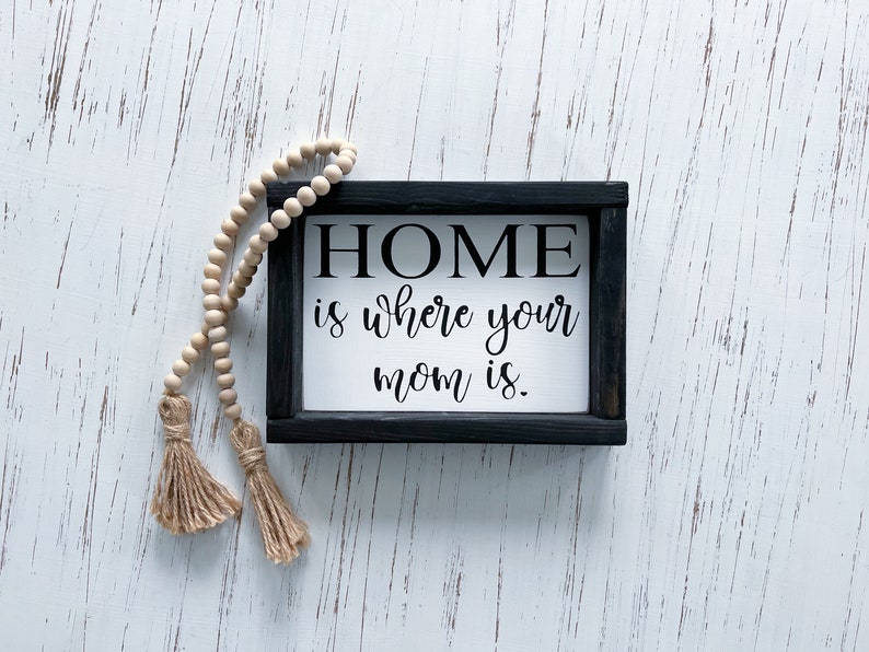 May include: A black framed sign with white background and the text "HOME is where your mom is." The sign is on a white wood background with a wooden bead garland with tassels.