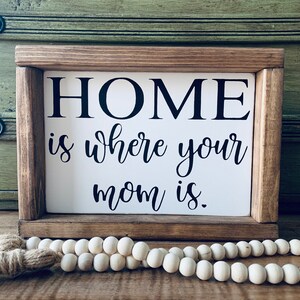 May include: A wooden sign with the text "HOME is where your mom is." The sign is framed in a rustic brown wood frame and has a white background.