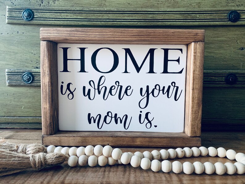 May include: A wooden sign with the text "HOME is where your mom is." The sign is framed in a rustic brown wood frame and has a white background.
